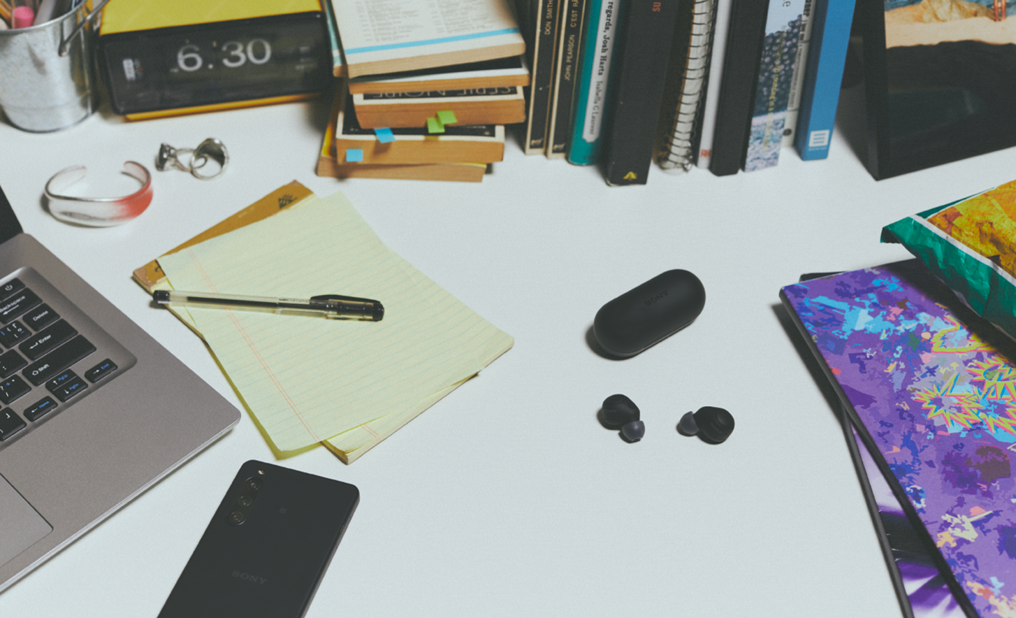 Image of black WF-C700N headphones and case on a desk with a notebook, laptop and mobile phone