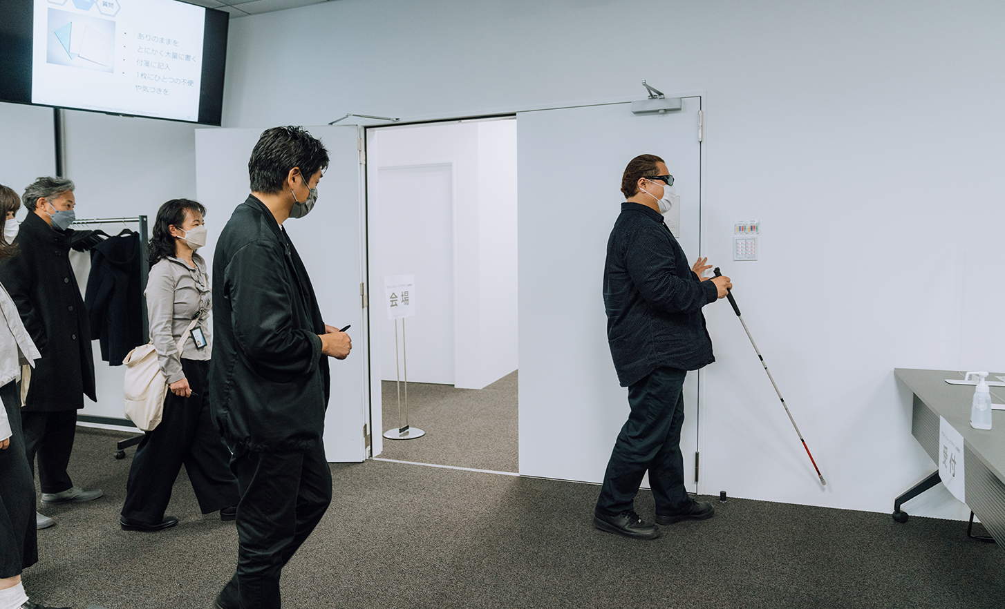 Des employés regardent l'utilisateur pilote avec sa canne blanche marcher dans la salle de conférence