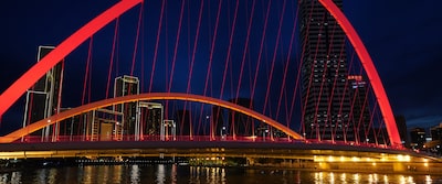 Nighttime image of a cityscape, with an illuminated red-coloured bridge in the foreground